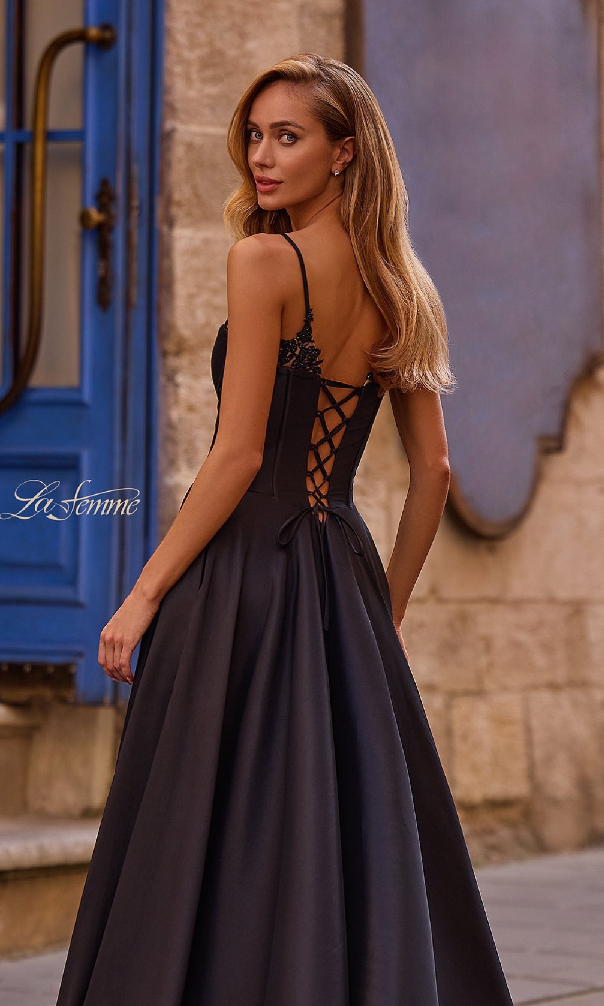 A woman in the La Femme 32878 Lace-Up Long Mikado Prom Dress, featuring a black formal design and lace-up back, stands outdoors on a stone street, gracefully looking over her shoulder.